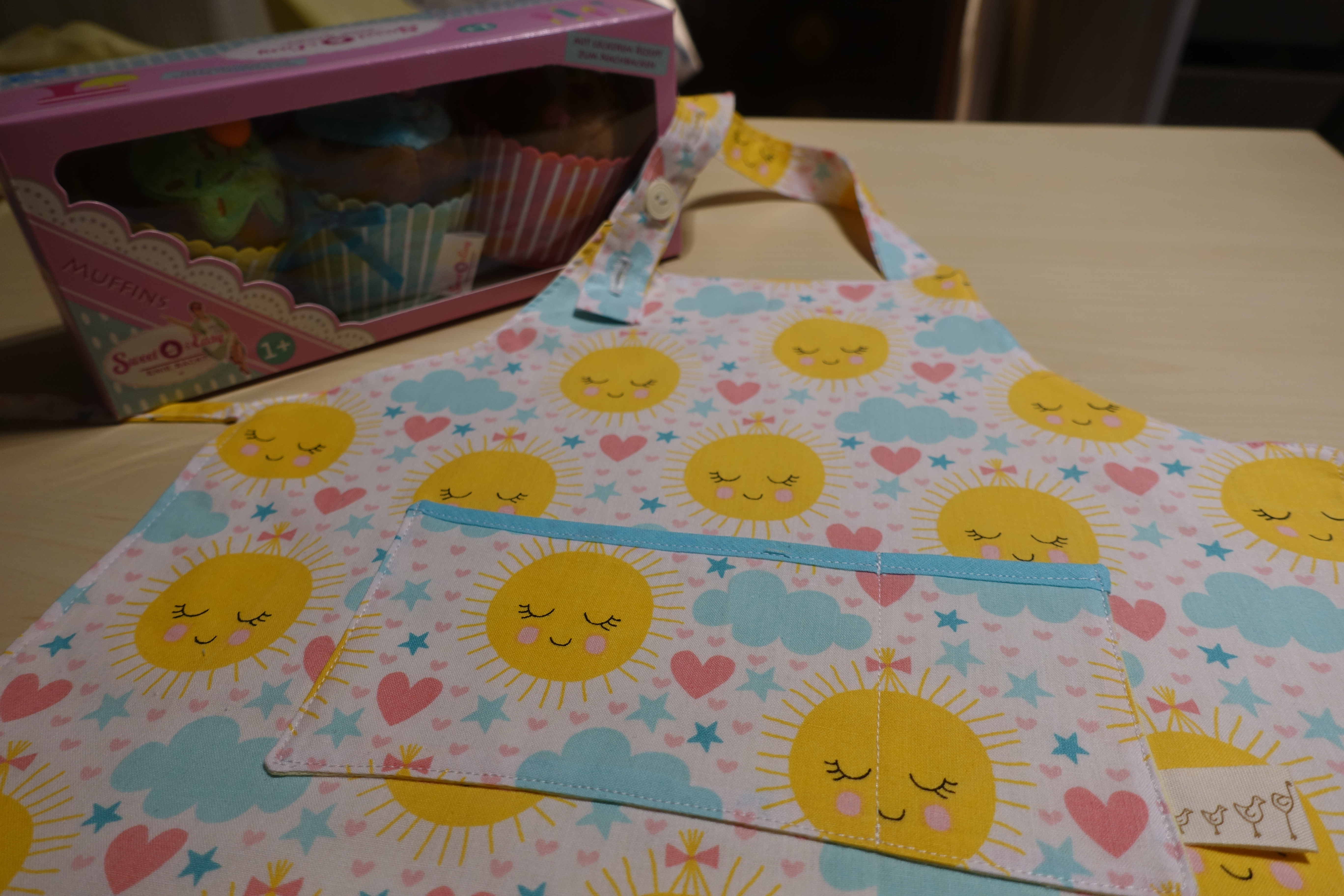 Handmade children's apron featuring a cheerful pattern of smiling suns, clouds, stars, and pink hearts. The apron includes a front pocket with light blue trim and is displayed beside a box of colorful cupcakes, highlighting its baking and crafting theme.